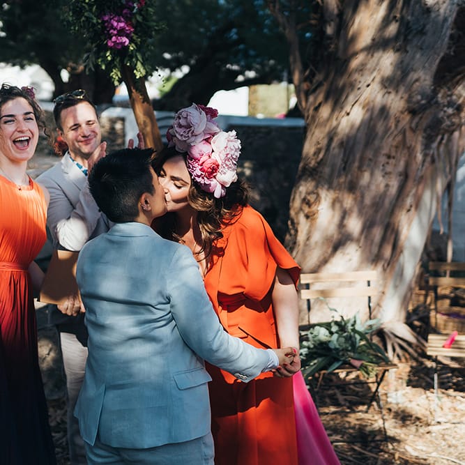 Brides kiss outdoors on their wedding day as wedding guests celebrate beside them