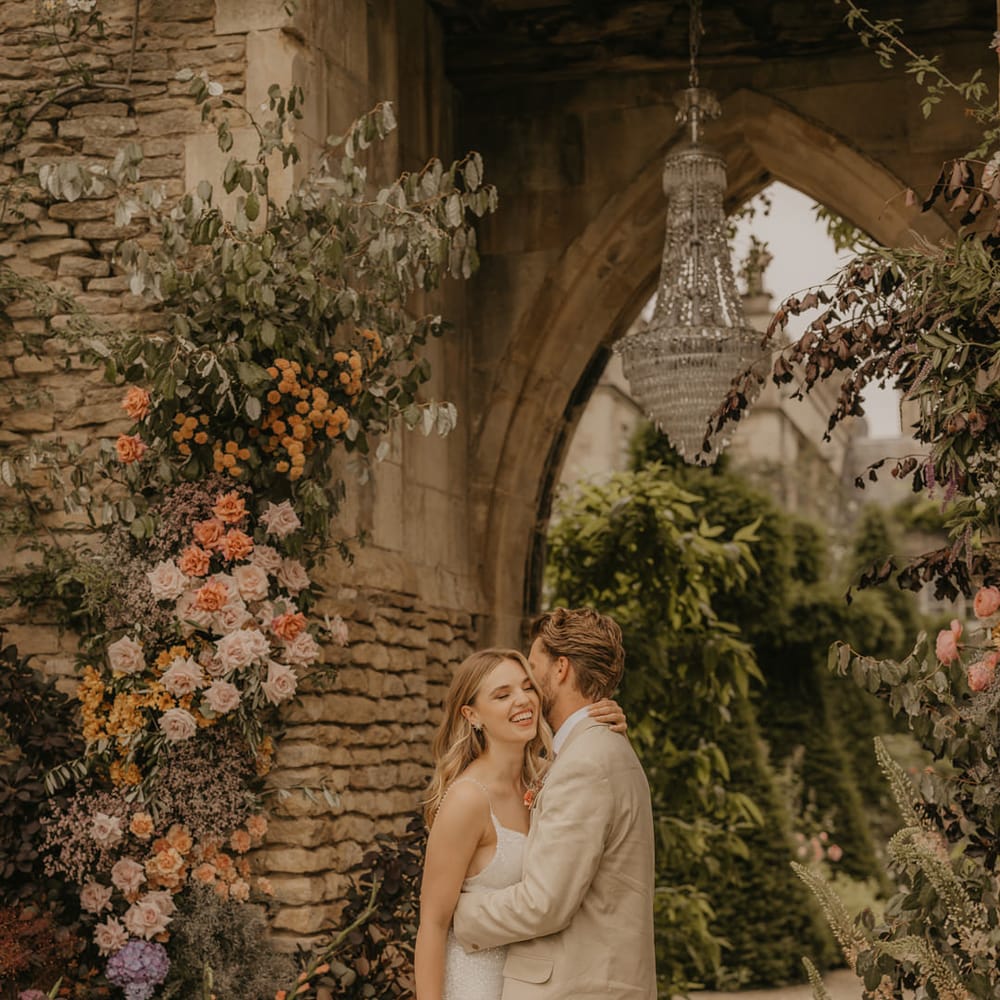 Groom in a beige suit embracing his bride in an embellished wedding dress by a pastel flower arrangement decorating the castle arch at Euridge Manor 