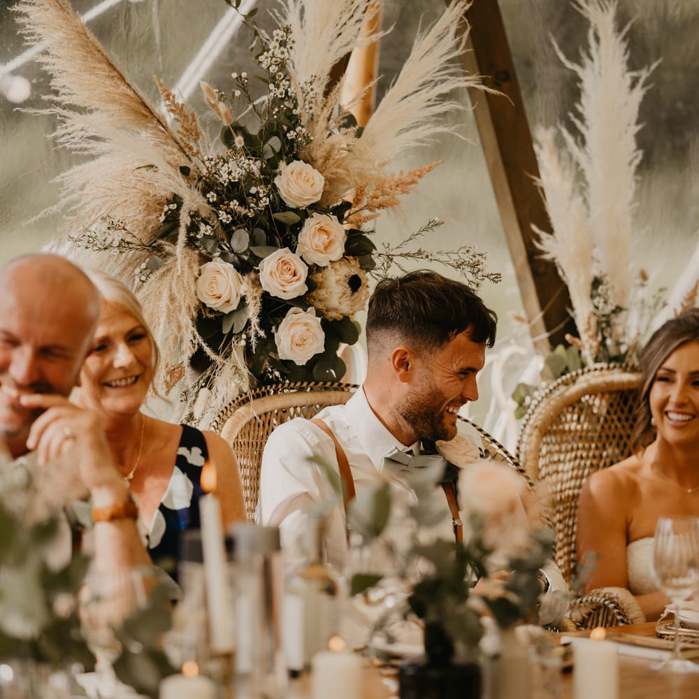 Bride & groom laugh during reception surrounded by pampas grass and sitting in wicker chairs | Mark Bamforth Photography