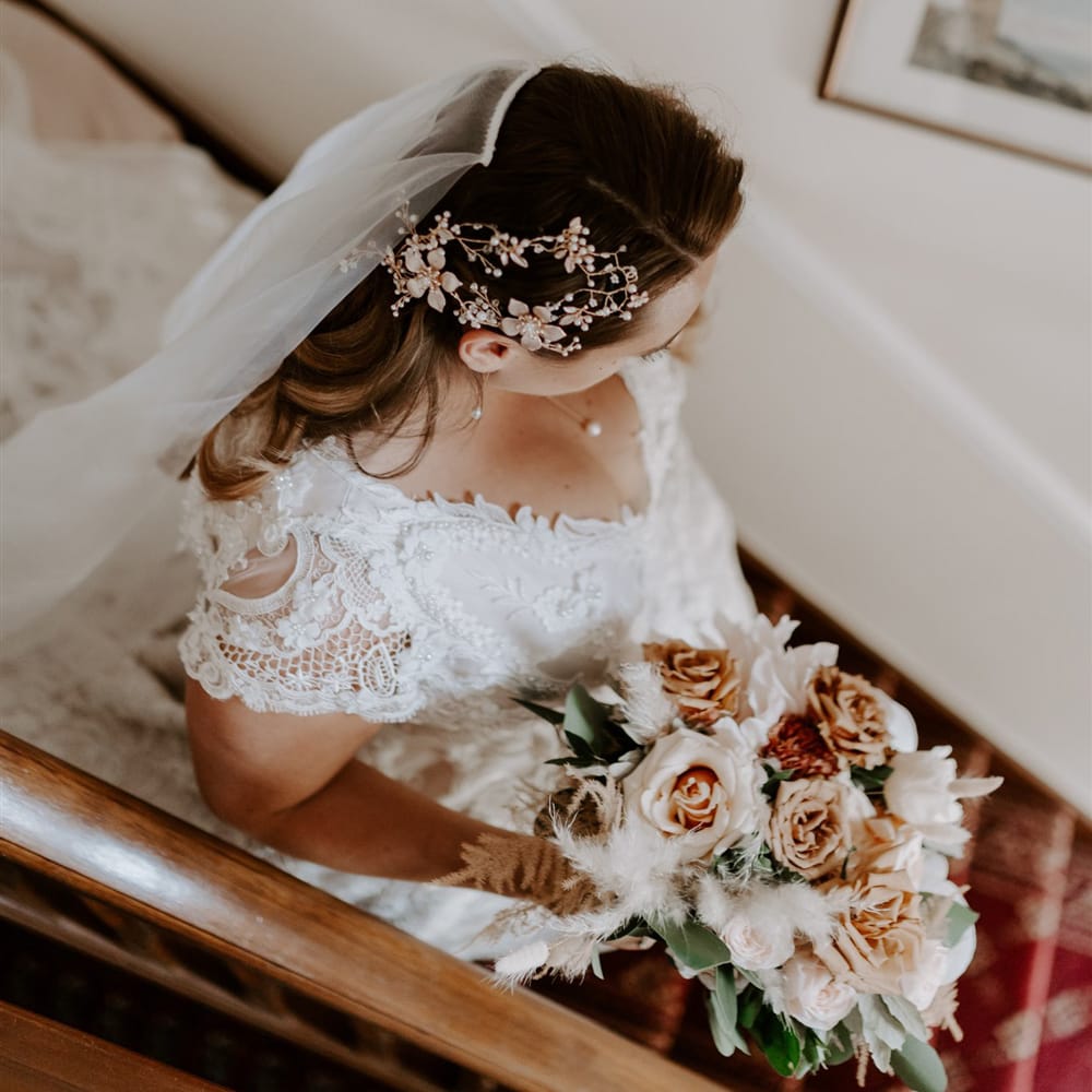 Bride in handmade wedding dress, veil and intricate bridal headpiece walks downs stairs holding mixed rose bridal bouquet before DIY garden wedding in Bedfordshire