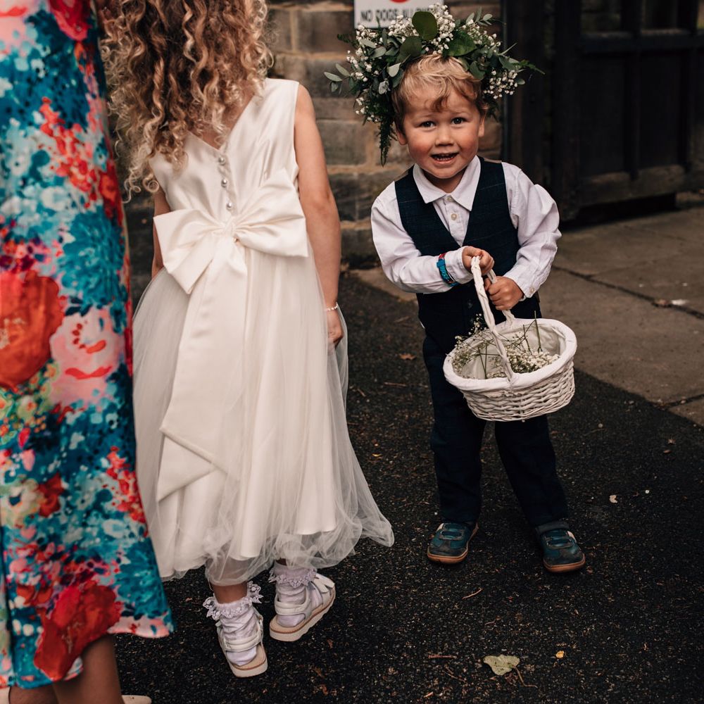 Flower boy in waistcoat and flower crown holds white basket with flower in before church wedding ceremony