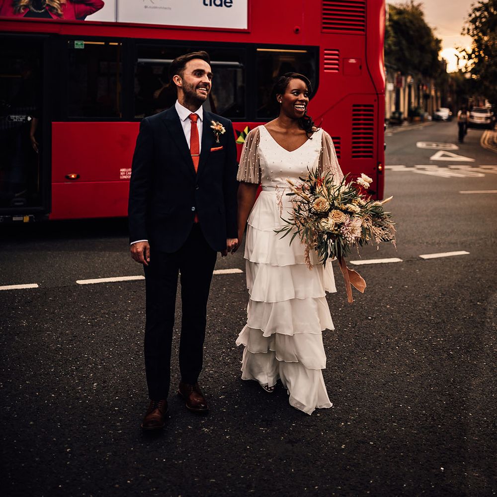 Black bride in a layered skirt and pearl top bridal separates holding hands with her groom in a navy suit in front of a red double decker London bus 