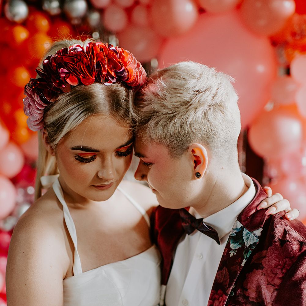 Bride with a ribbon headdress, and long eyelashes embracing her bride in a floral velvet suit jacket at retro wedding 