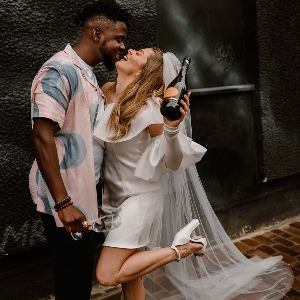 bride in a short wedding dress holding a Prosecco bottle with her groom in a patterned shirt at their city elopement 