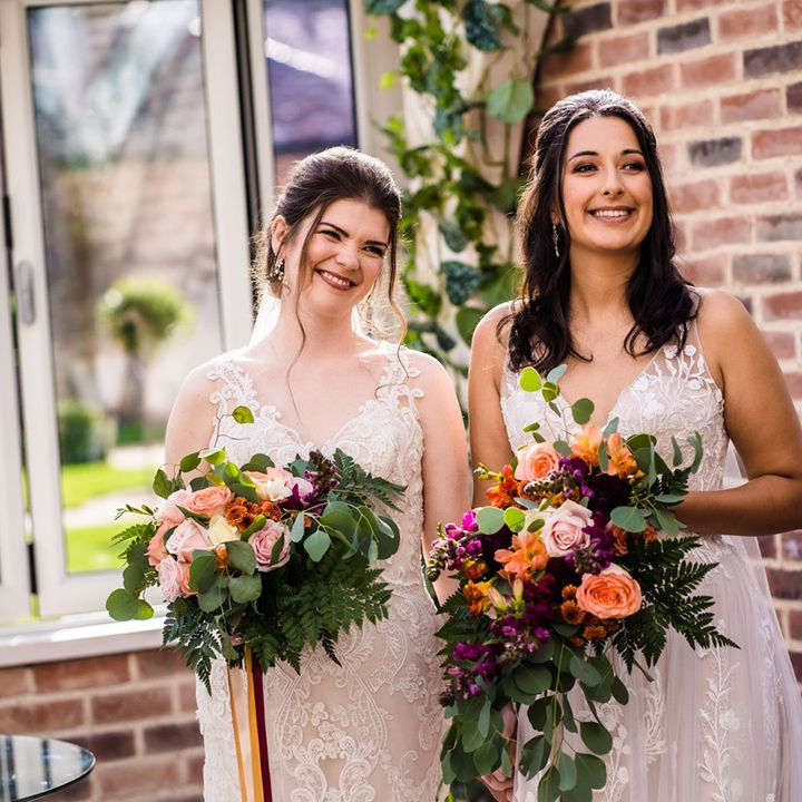 Brides stand together holding purple, pink, and orange flowers with roses and fern leaves