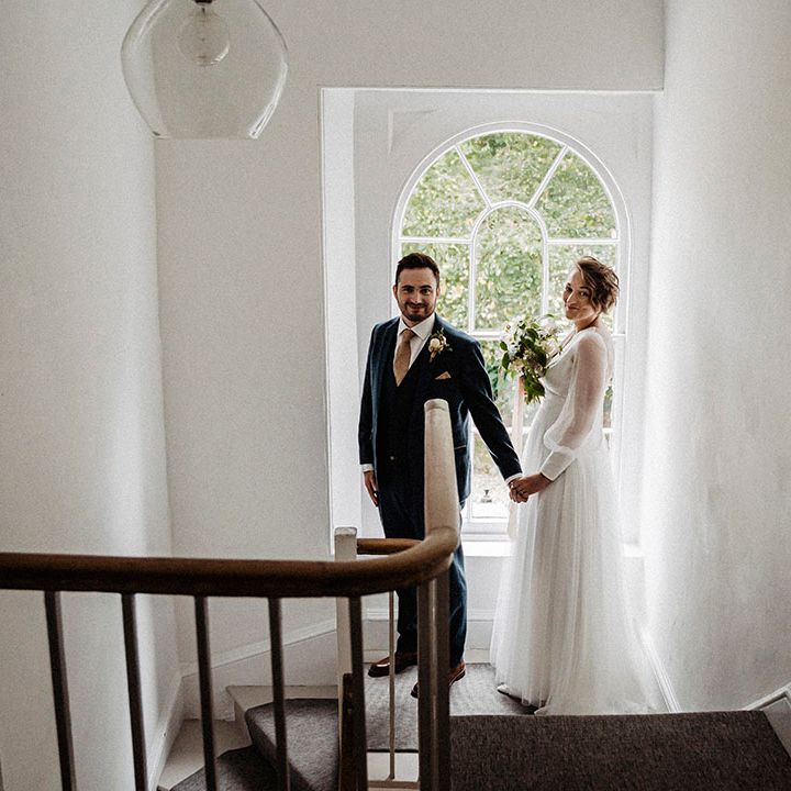 Bride in sheer long sleeve wedding dress with tulle skirt standing with groom in blue suit with gold patterned tie