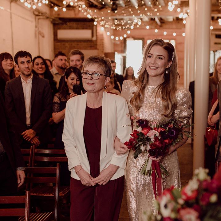 Mother of the bride in burgundy dress and white blazer walks the bride in a gold wedding dress down the aisle 