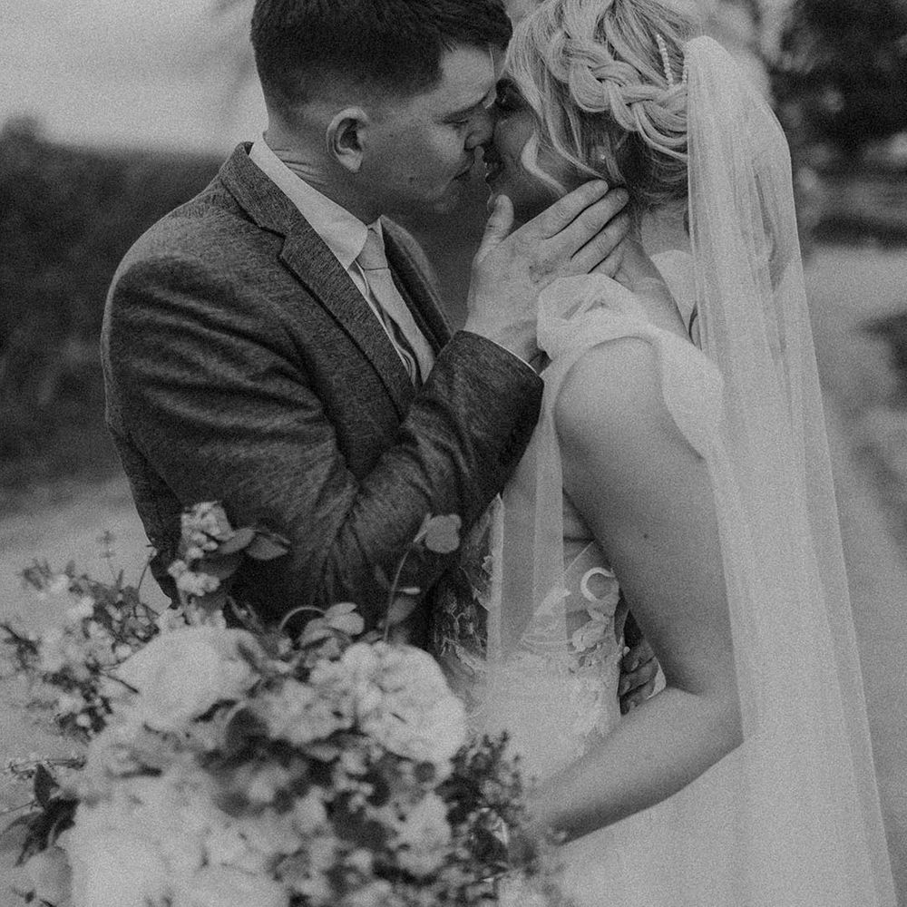 Bride with blonde hair in braided ponytail with diamanté hair clip kissing groom holding wedding bouquet
