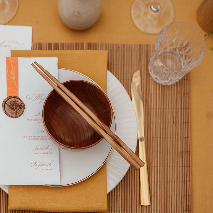 Place setting with yellow table rubber, bamboo place mat, gold cutlery, brown rice bowls, wooden chopsticks and menus card for Japanese wedding 