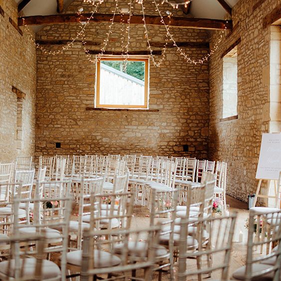 The Barn At Upcote wedding ceremony room complete with white wicker chairs and fairy lights hanging from above in rustic styled room