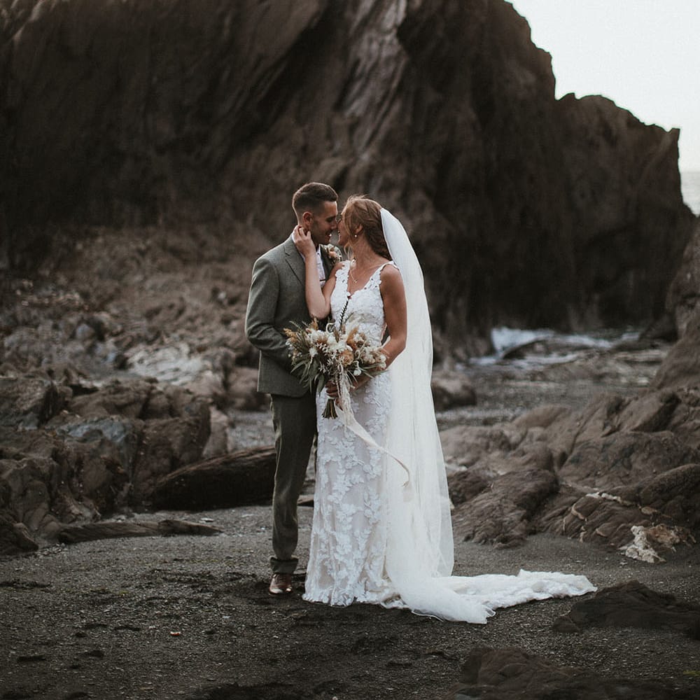 Bride holds neutral toned bridal bouquet tied with ribbon as she leans in to kiss her groom at Tunnels Beach