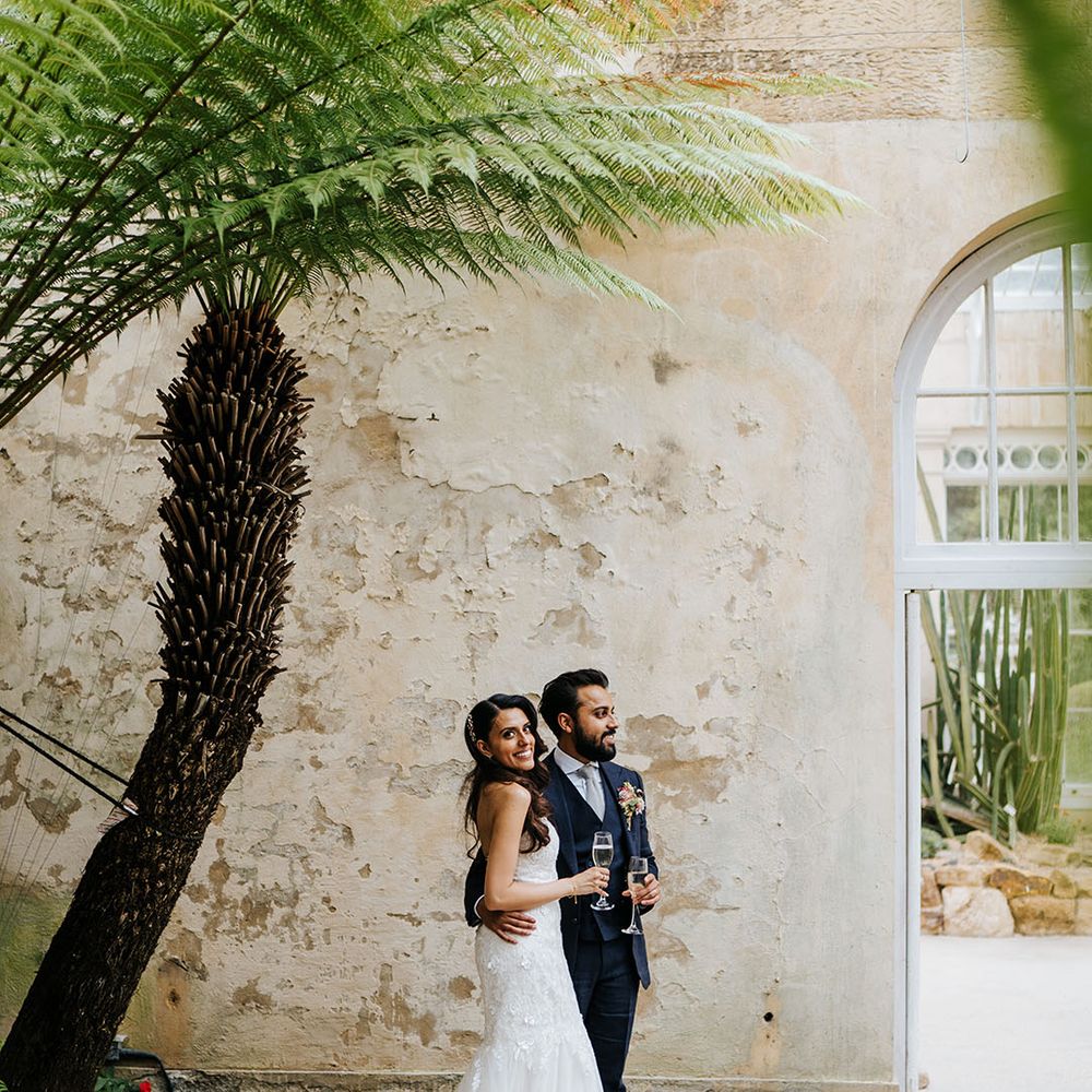 Bride & groom stand beside rustic wall at the Great Conservatory in Syon Park