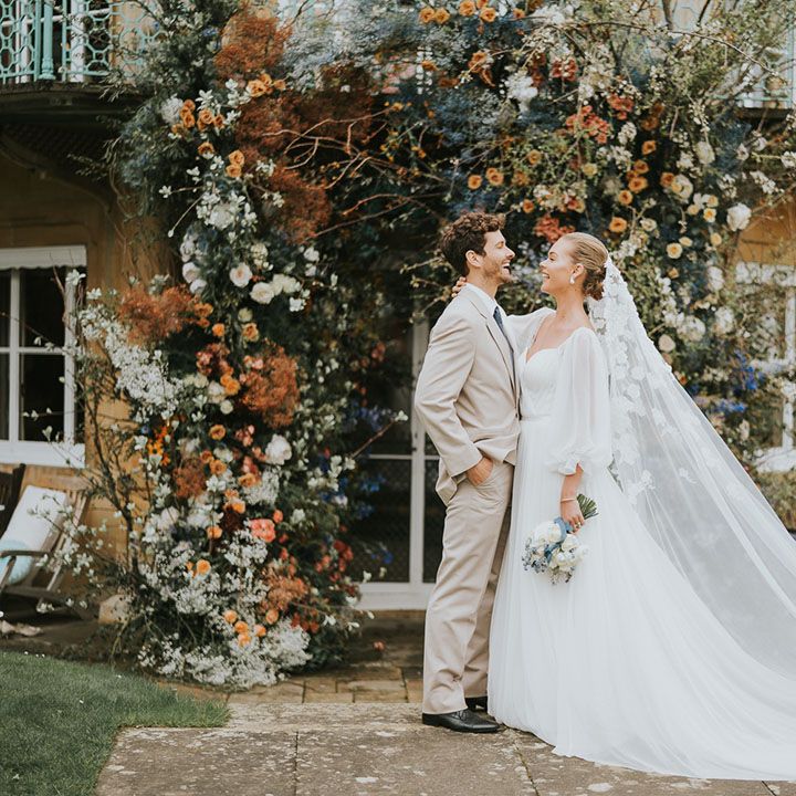 Bride and groom laughing together standing in front of blue and orange wedding flower arch 