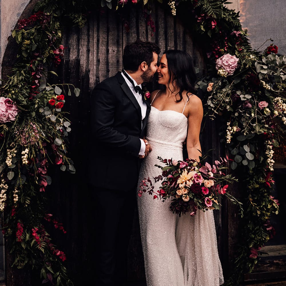 The bride in a sparkly wedding dress smiles with the groom in black tie under the pink, red and purple wedding flower arch 