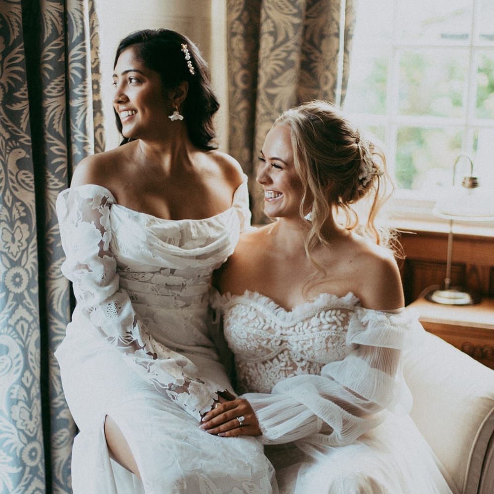 Bride in off the shoulder long sleeve wedding dress sitting on bride's lap wearing strapless lace wedding dress on cream coloured chair