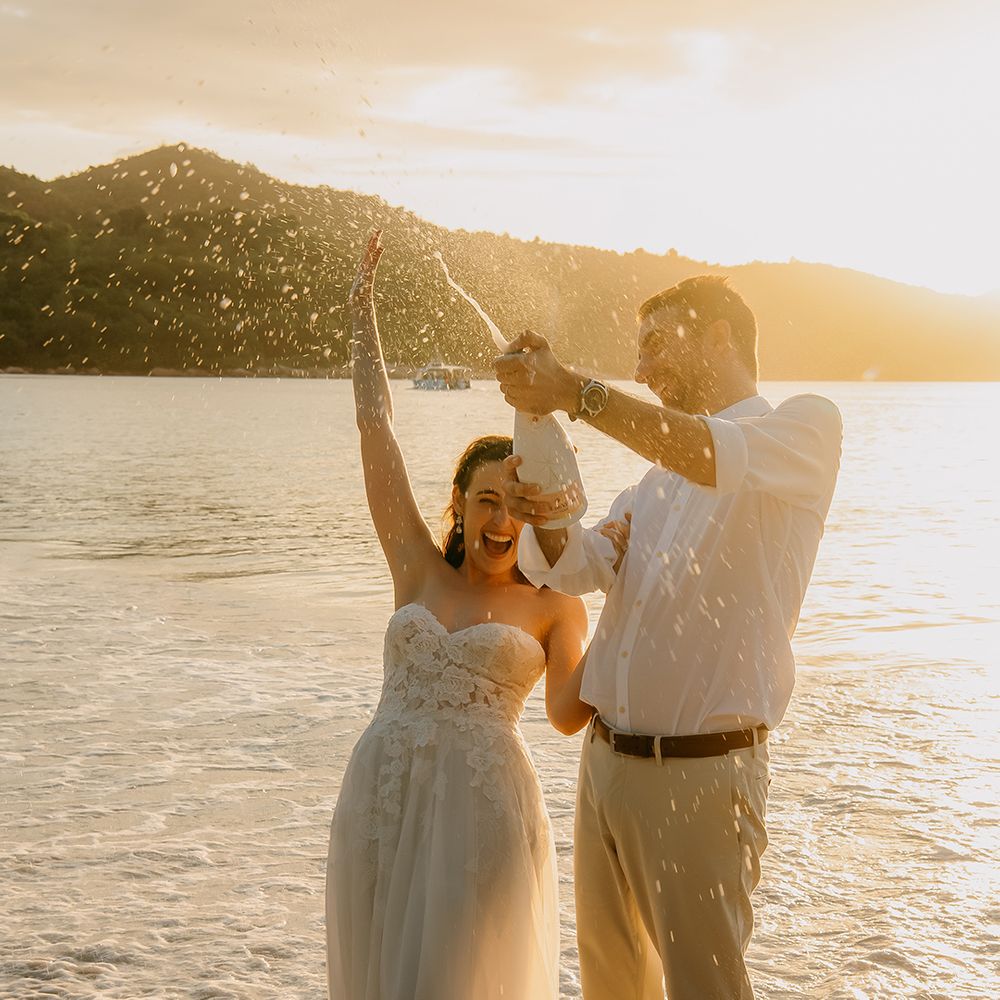 Bride in off the shoulder lace wedding dress with sweetheart neckline and lace floral details and groom in  crisp white shirt, light coloured chinos, tan leather belt and white trainers doing champagne pop on the beach