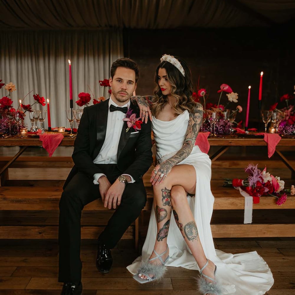 Groom in classic black tuxedo with magenta amaryllis and orchid boutonniere and bride in cowl neck sleeveless satin wedding dress with leg slit, gold glittery wedding headband, and soft grey fluffy feather open toe strappy high heels sitting on steps of Elmore Court with red tapered candles, dark brass candlestick holders, red roses and pink peonies decorations