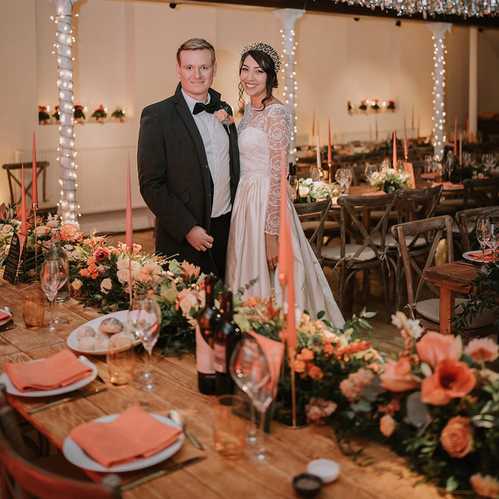 Bride in taffeta material princess wedding dress with long lace sleeves  and groom in black tux standing in reception room with silver and gold streamer decorations and fairy lights 