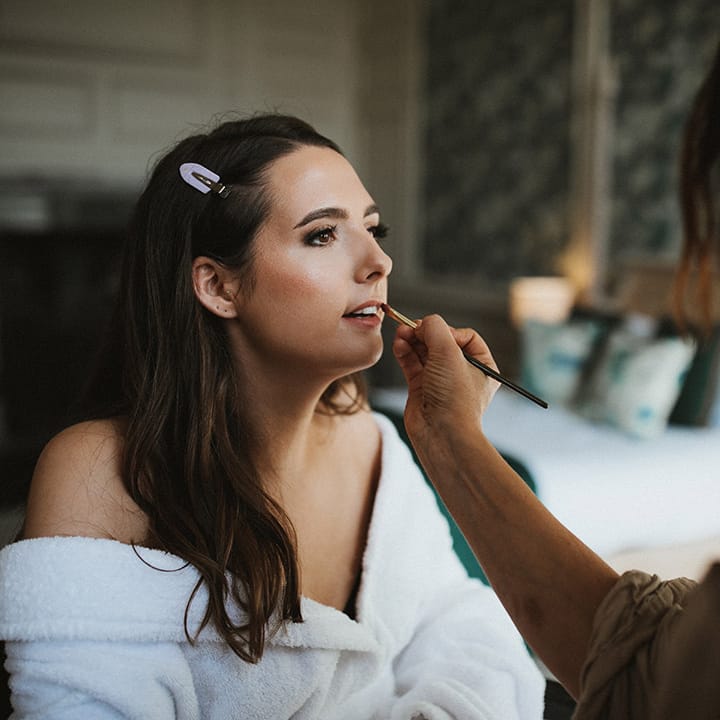 Bride sits in a white robs getting ready for the wedding day as she has her makeup done 