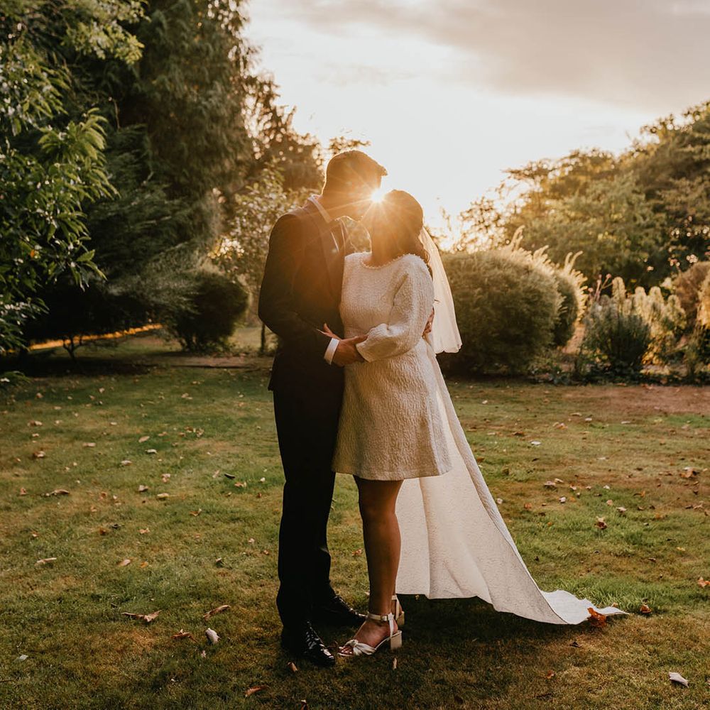 The groom in black tuxedo kissing the bride during sunset golden hour with the bride in a short wedding dress with train and bow detail and shoulder length veil