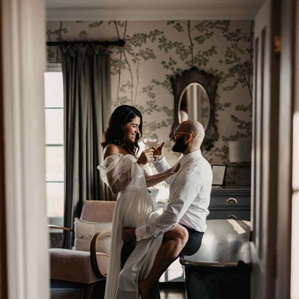 Bride in sheer puff sleeve wedding dress with front slit embracing groom in white shirt and black suit trousers, sharing a drink together 