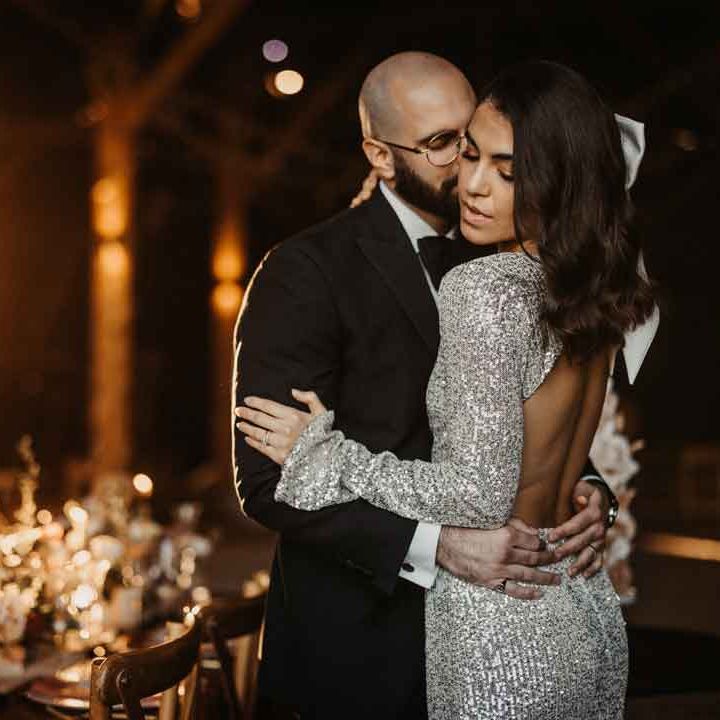 Bride in long sleeve silver sparkly wedding dress with open back detail embracing groom in classic black tuxedo 