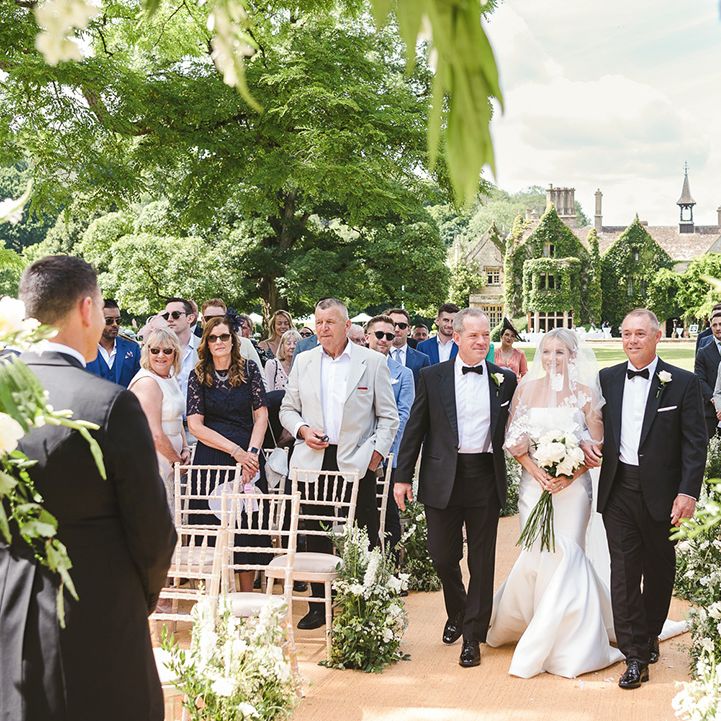 Two men in black tie walk the bride in a fitted wedding dress down the aisle for the outdoor wedding ceremony 