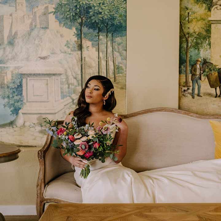 Bride in bow front sleeveless satin wedding dress with large gold bridal hair clip sitting on cream and gold chaise lounge holding large dried flower, eucalyptus, spring florals and garden rose bridal bouquet 