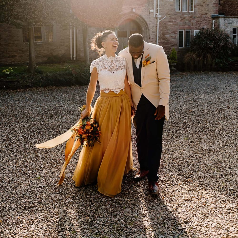Groom in cream morning suit with forest green grooms waistcoat and trousers, forest green pocket square and orange boutonniere walking with bride in white mesh lace bridal crop top and yellow wedding skirt holding dried autumnal wedding bouquet with sunflowers, garden roses, pampas grass, eucalyptus, yellow tall kangaroo paws, orange osmanthus flowers, foliage and dried flowers at Broadfield Court