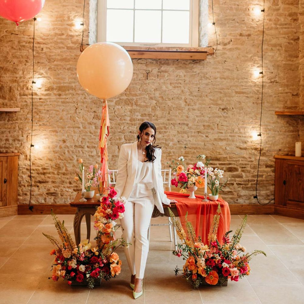 Bride in white bridal suit with closed toe bright green wedding heels standing by metallic pink, white, orange and yellow balloons, wedding fairy lights decorations and wooden table with orange table runner and mixed seasonal flower arrangements 