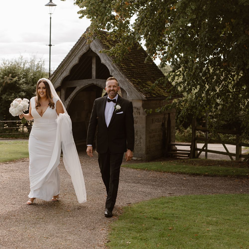 Bride in white Made With Love wedding dress and white chapel length veil holding white and blush pink rose bouquet whilst walking through churchyard with father in black tuxedo