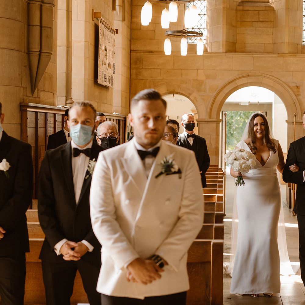 Smiling bride in Made With Love wedding dress walks arm in arm with father in black tuxedo up the church aisle towards groom in white double breasted tuxedo jacket