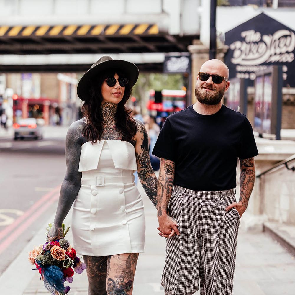 Bride in an alternative wedding dress, bridal boots and fedora hat holding hands with her groom in a check suit and black t-shirt 