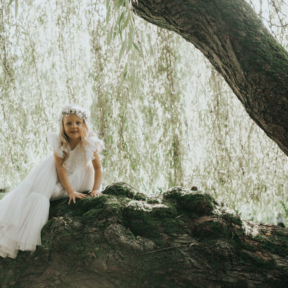 A flower girl in a long white dress and flower crown sits atop a rock at Crab & Lobster fairytale wedding. 