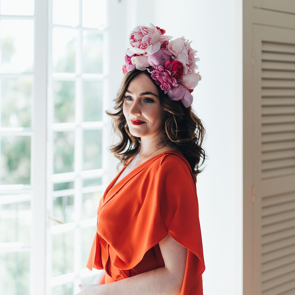 Bride wears red gown with pink bridal crown as she stand beside window on her wedding day in Menorca