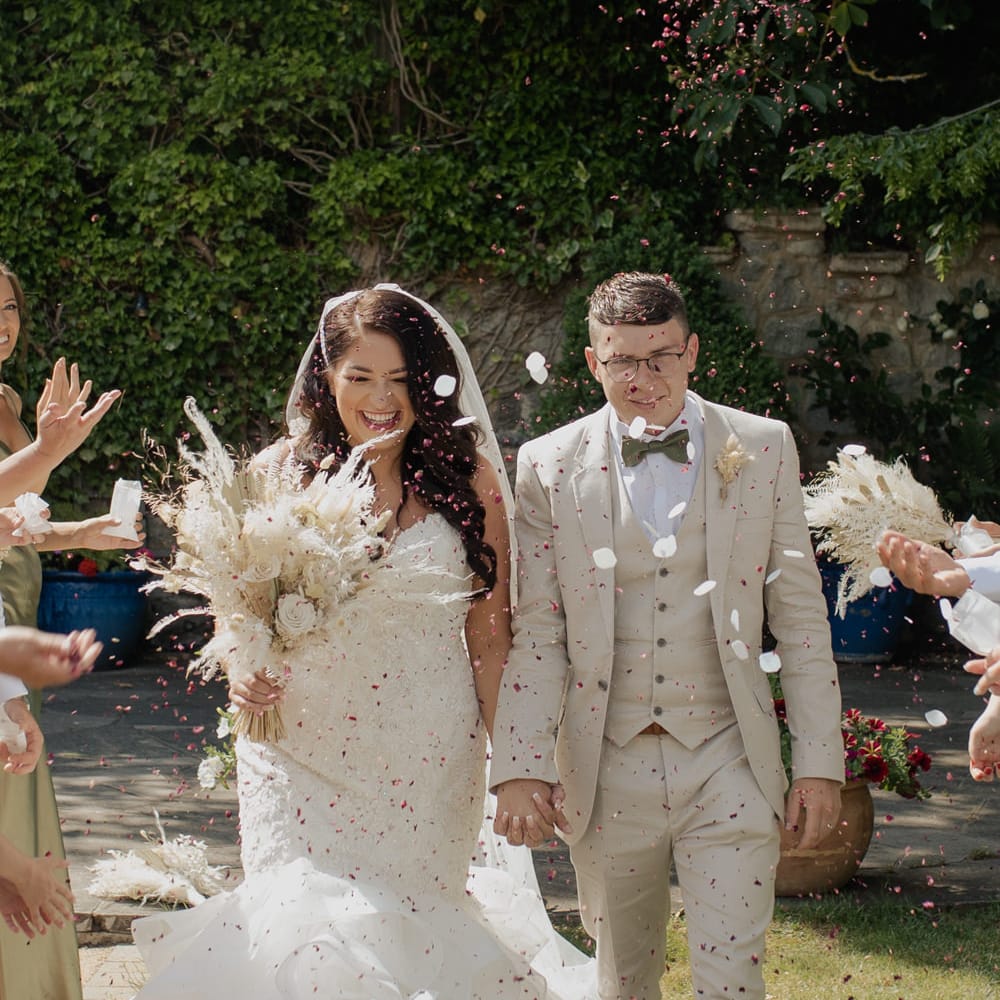 Confetti moment with groom in a beige suit holding hands with his bride in a Mori Lee fishtail wedding dress