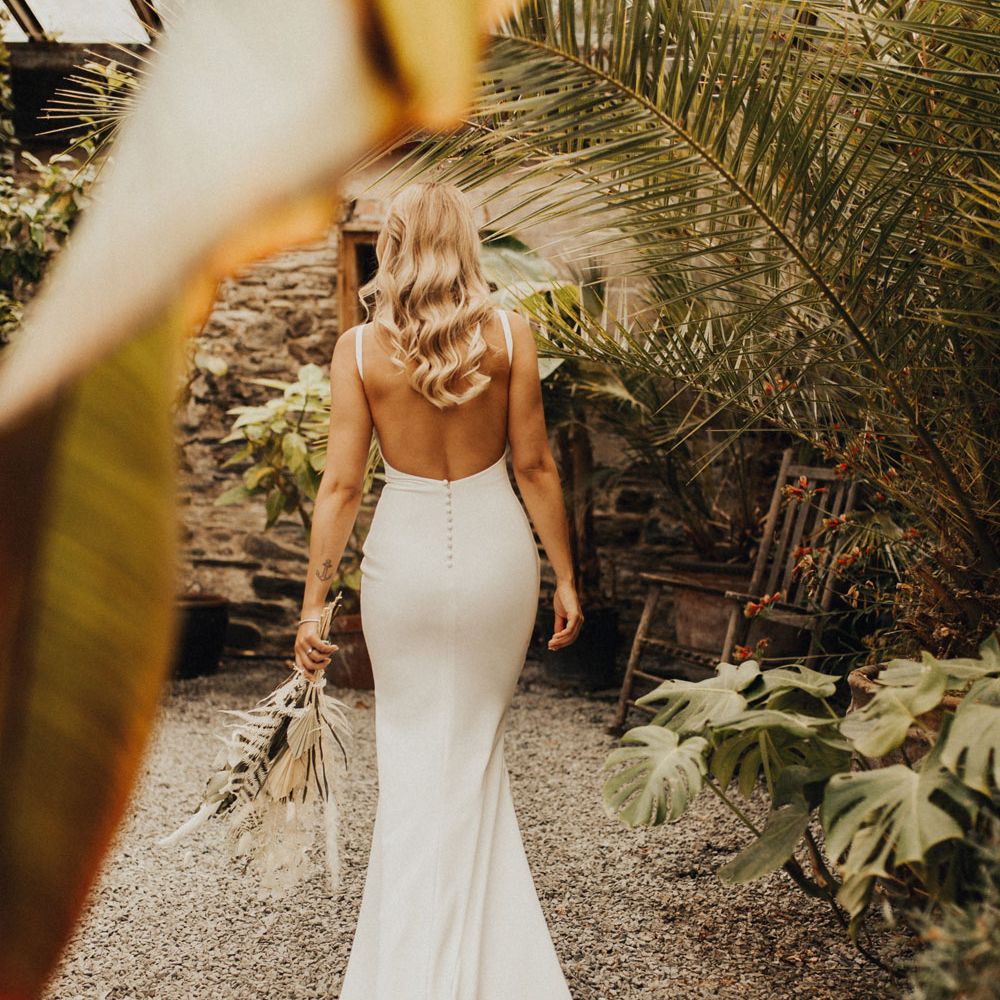 Bride in white open back Made With Love wedding dress with button detailing holding white and green dried bridal bouquet stands in glasshouse at Anran Devon