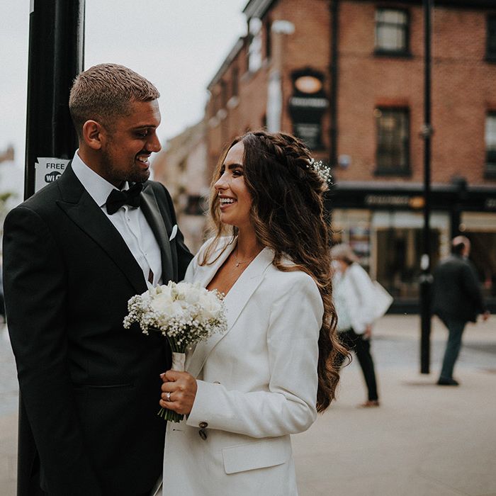 Bride in a white three piece suit holding a white flower bouquet by a lamppost with her groom in a tuxedo 