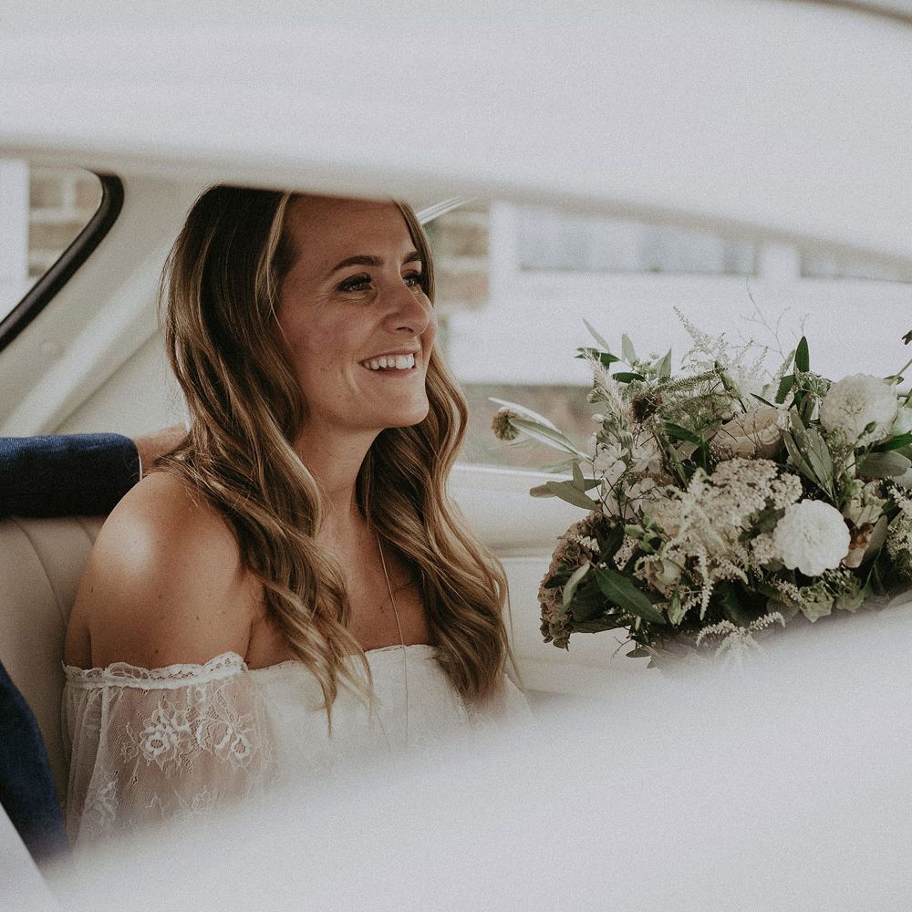 Bride with curled hair wearing Grace Loves Lace bardot wedding dress holding white and green bridal bouquet sits in back of VW bug before Isle of Wight wedding with macrame wedding decor