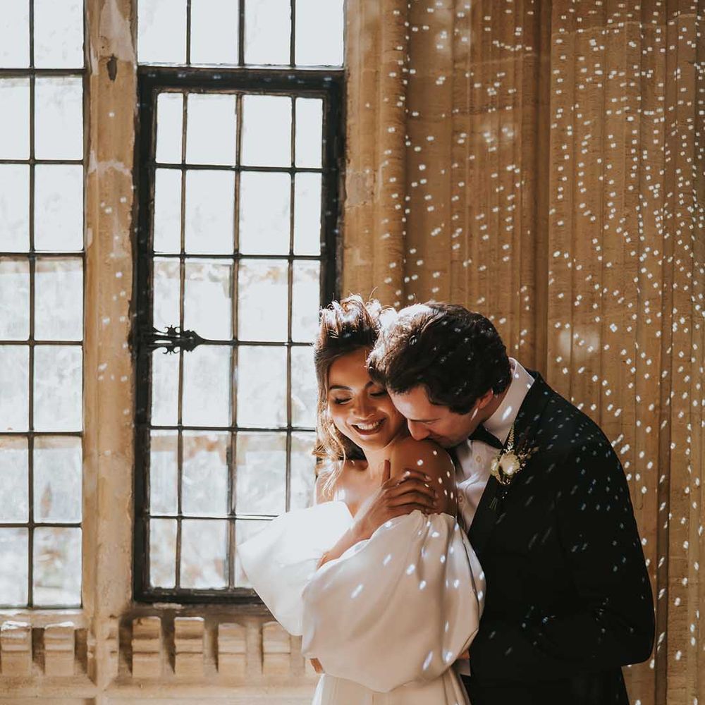Groom in a tuxedo kissing his brides shoulder in a strapless wedding dress with glistening lights all around them 