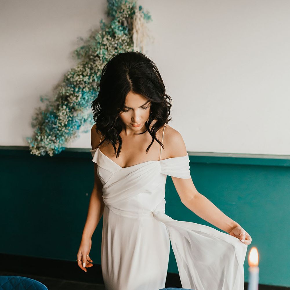 Bride in a cold shoulder wedding dress standing next to plush teal velvet reception chairs 