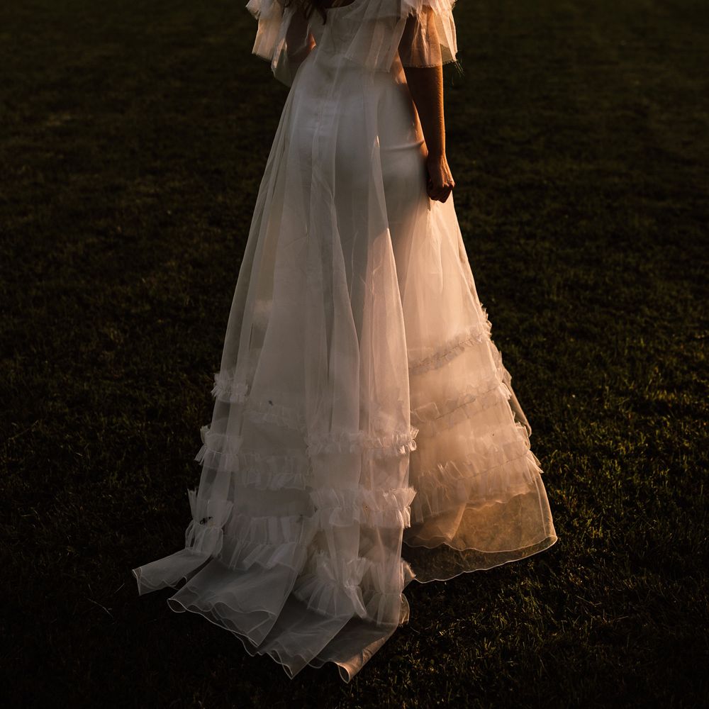 Bride in white Halfpenny London Mayfair dress with riddle sleeves stands in garden during golden hour at Cornish garden wedding reception 