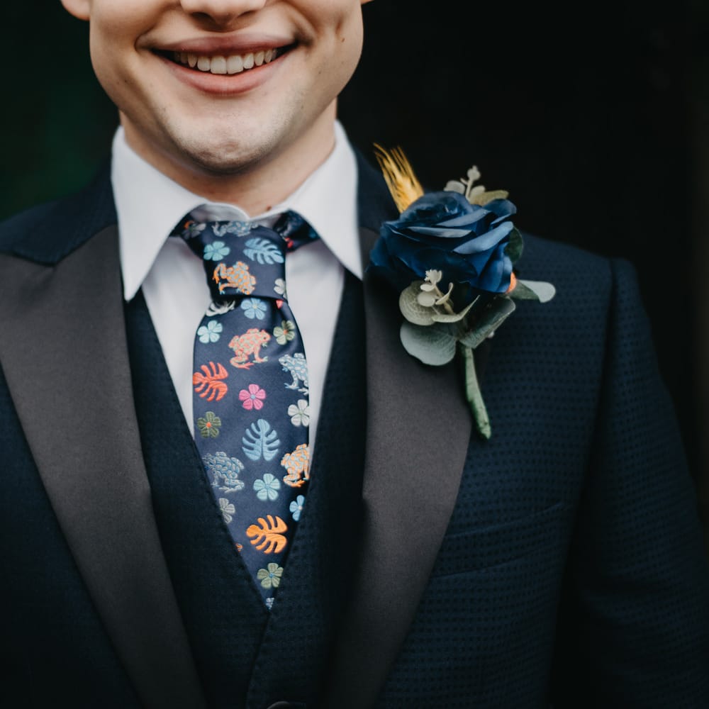 Smiling groom in dark blue suit, white shirt, blue florals tie and blue rose buttonhole for The Wellbeing Farm wedding 