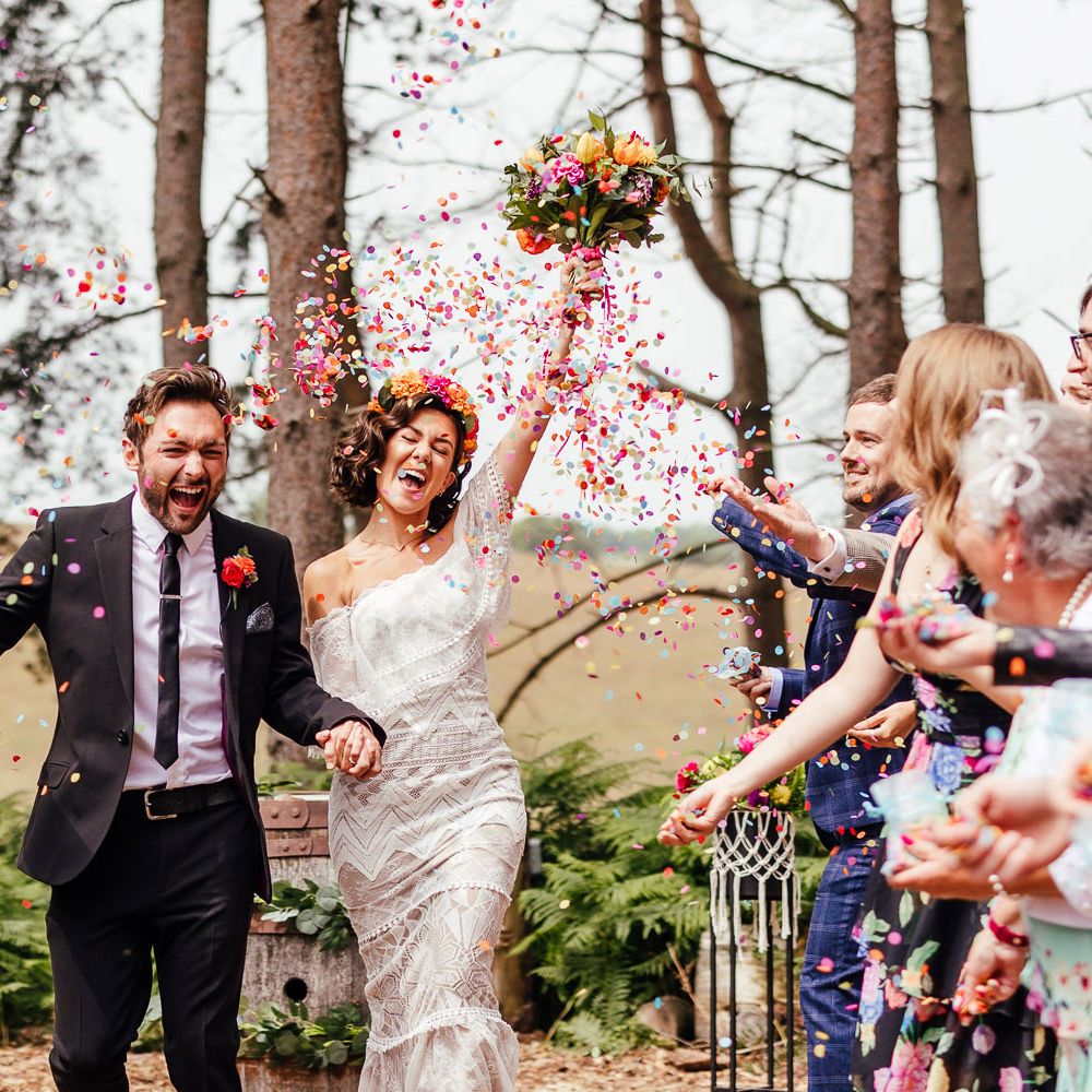 Bride in off the shoulder Grace Loves Lace wedding dress and bridal flower crown holds colourful wedding bouquet and the hand of groom in black suit as guests throw multicoloured confetti