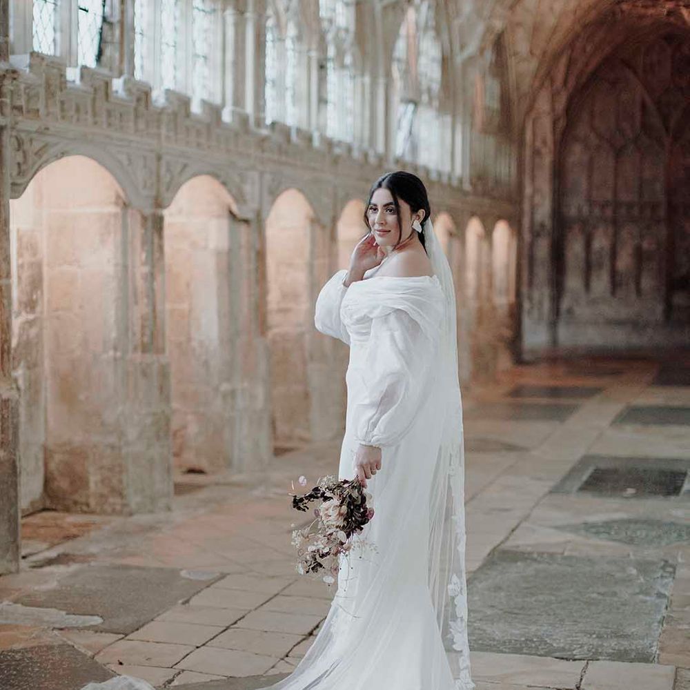 Bride with sleek back hair in an off the shoulder wedding dress with long sleeves and a cathedral length veil with appliqué edge 