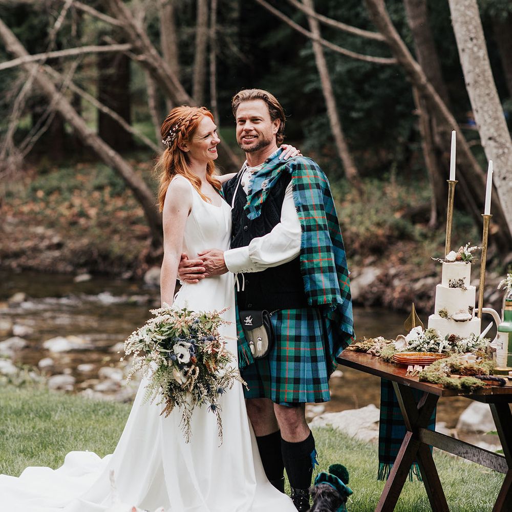 Groom in a kilt and bride in an a line wedding dress standing next their dessert table with their Scotty dogs by their feet