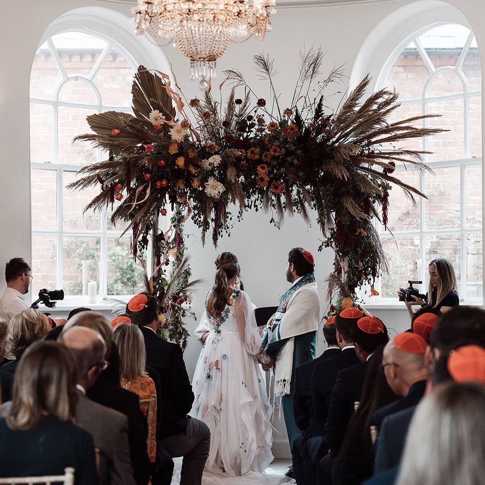 Bride in long mesh sleeve wedding dress with floral embroidery stands with groom in kippah and green suit under arch with dried flower cloud for interfaith wedding ceremony
