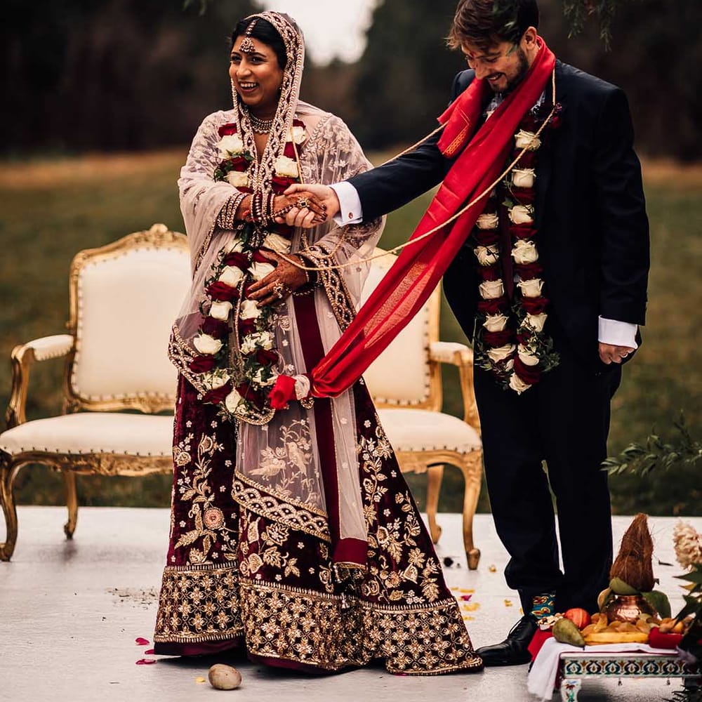 Bride wears Indian burgundy saree wedding dress and holds hands with groom who wears matching tie 
