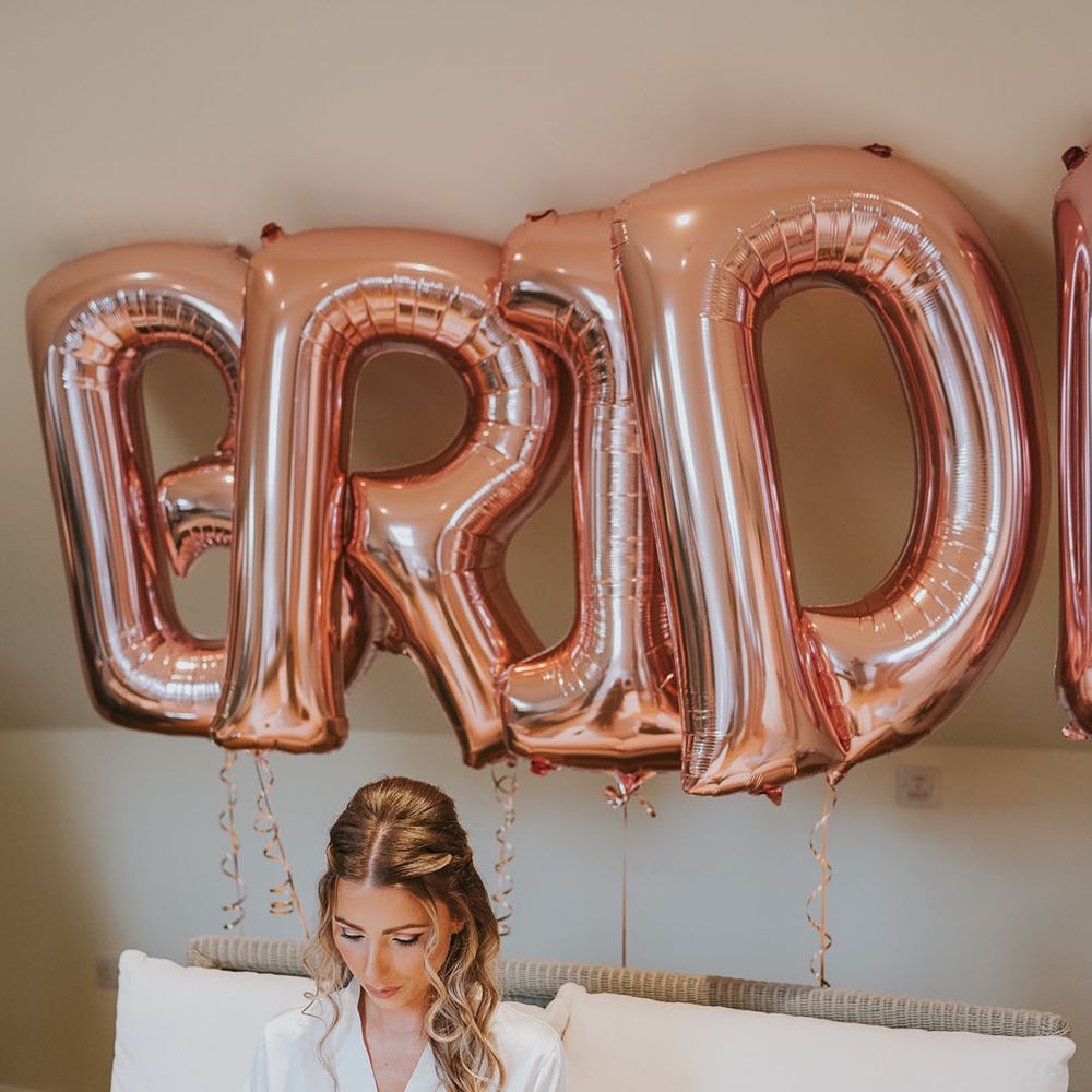 Bride in satin dressing gown reads letter and opens gift from the groom in front of rose gold 'bride' balloons
