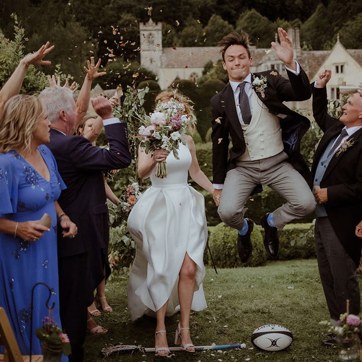 Groom jumps for joy over a rugby ball with bride as guests throw confetti in congratulations 